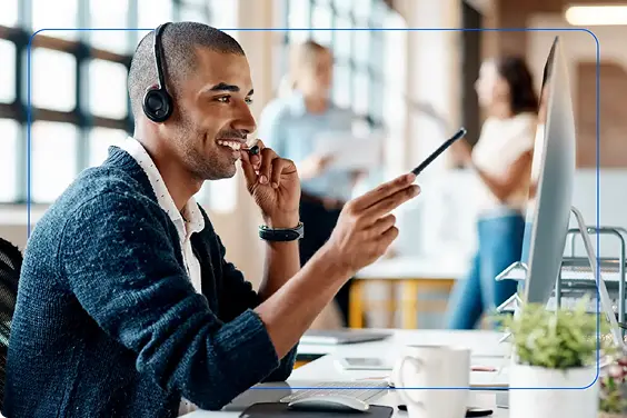 A smiling man caling with Headphones on a desk with a man and woman talking in background
